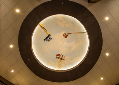 Ceiling view of the High Reach lobby that shows lighting and a painted sky with commercial construction equipment.